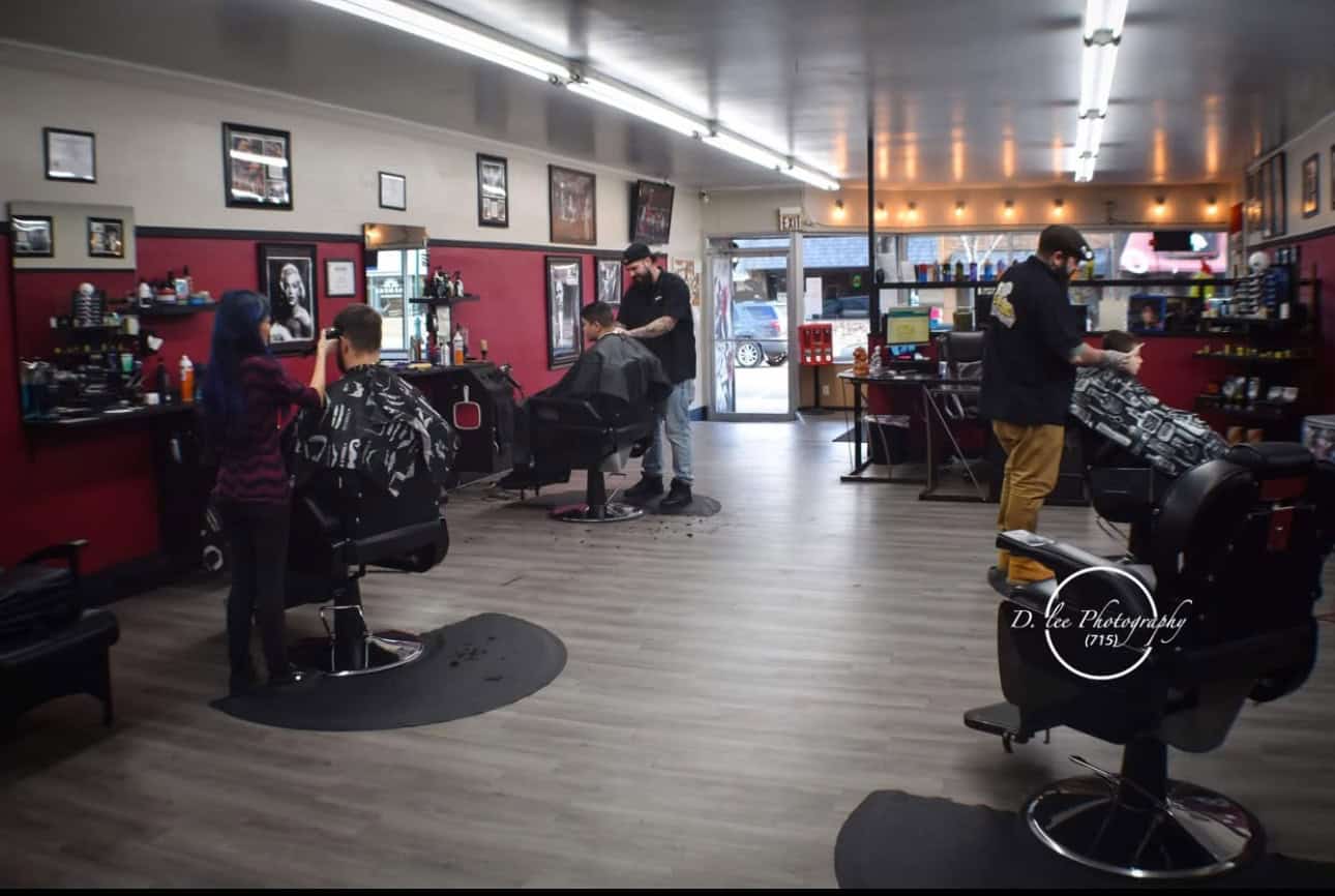 Inside The Shawano Barbers: three of the shop's barbers mid-cut, clients caped, under the red walls and framed portraits, on the wood floor.
